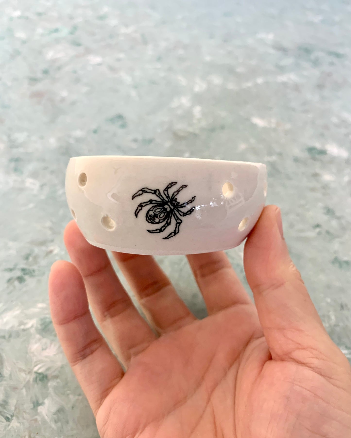 A hand holding a white porcelain tealight candle holder with a black spider design.