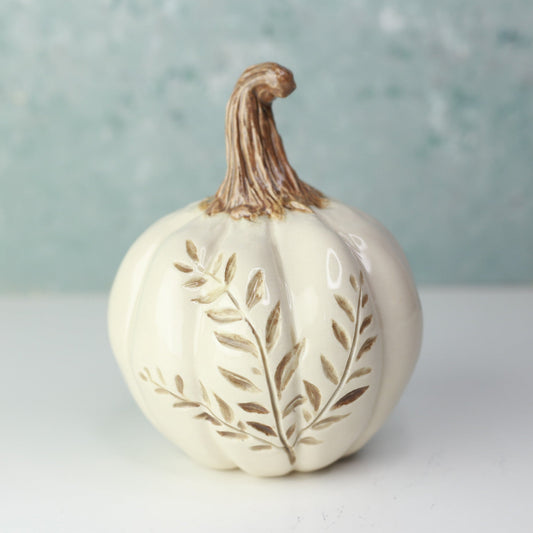 An off-white ceramic decorative pumpkin with a brown stem and embossed leaf details, set against a soft, textured light blue-green background.
