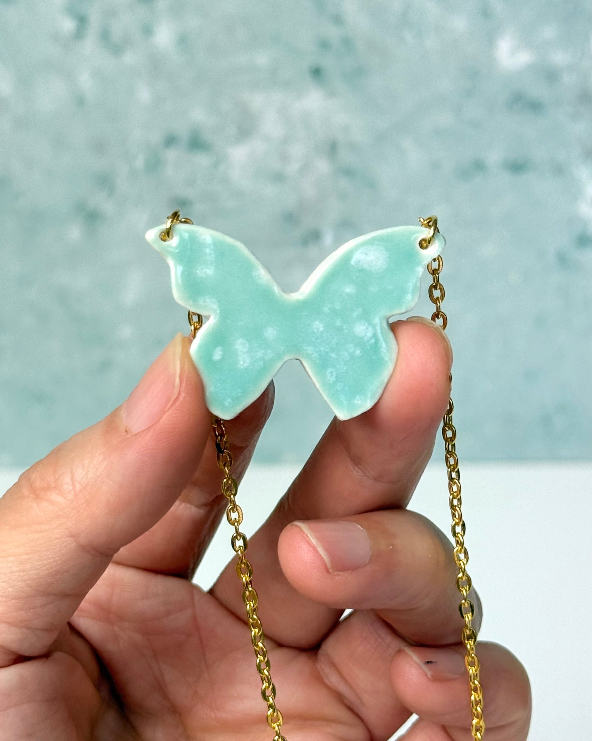 Butterfly-shaped pendant with gold chain held by a hand against a light blue background