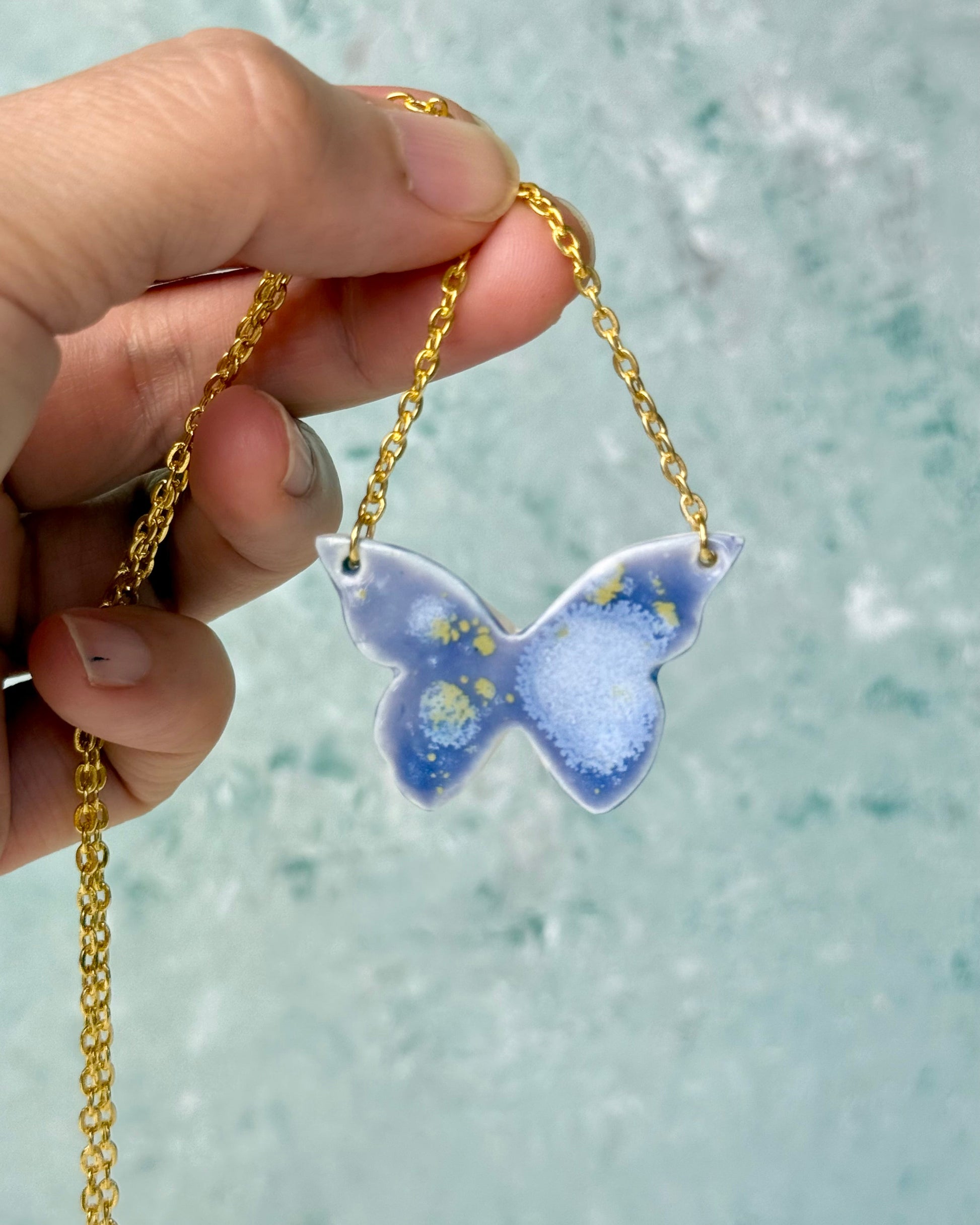Butterfly-shaped pendant on a gold chain held by a hand against a blurred background