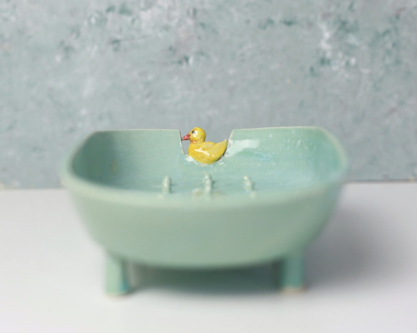 Small ceramic bathtub soap dish with a yellow duck toy on a textured gray background