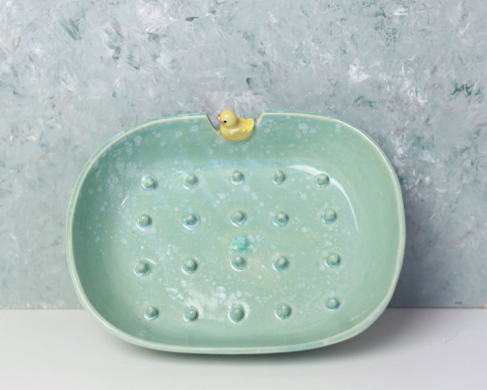 Green ceramic soap dish with a small yellow duck on a textured gray surface