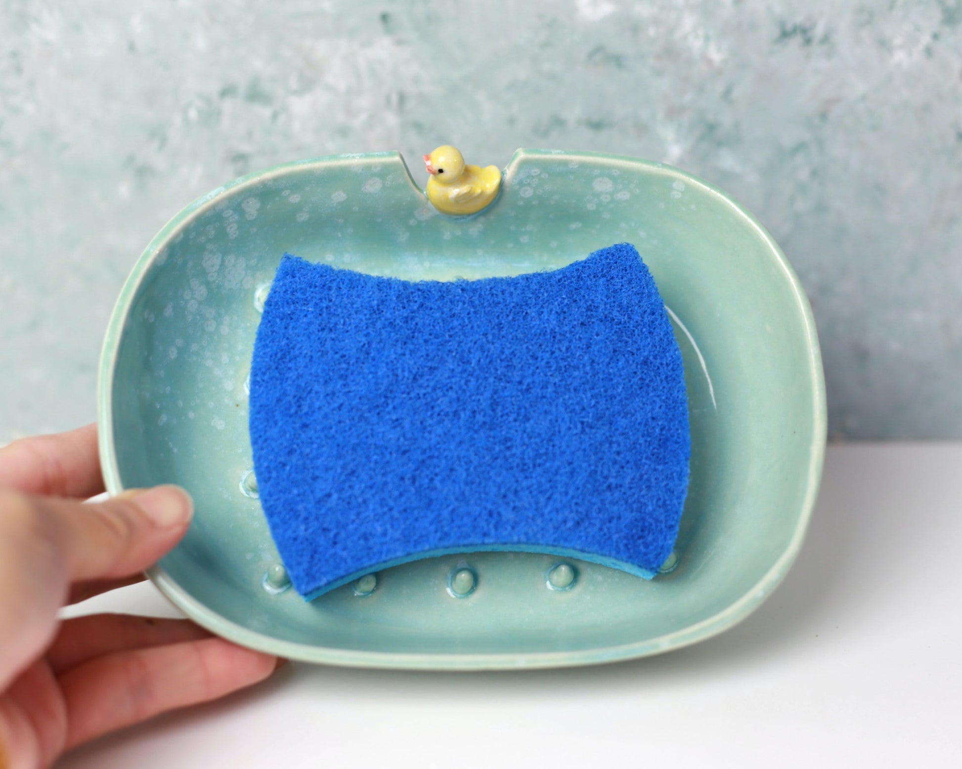Blue sponge on a green soap dish with a yellow rubber duck, held by a hand against a light gray background.