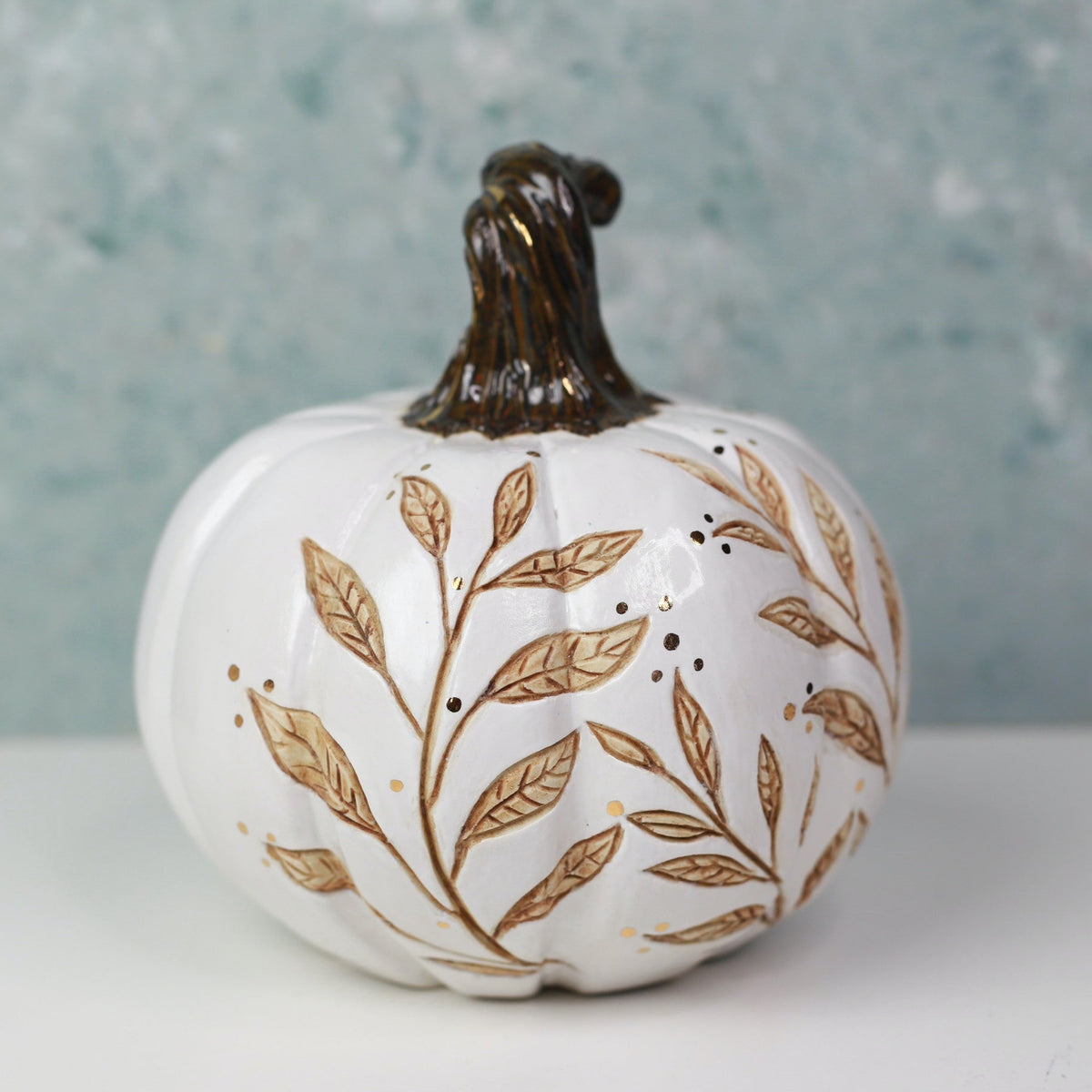 A white ceramic decorative pumpkin with a dark brown textured stem, decorated with gold-toned leaf and berry patterns, set against a textured light blue-gray background.