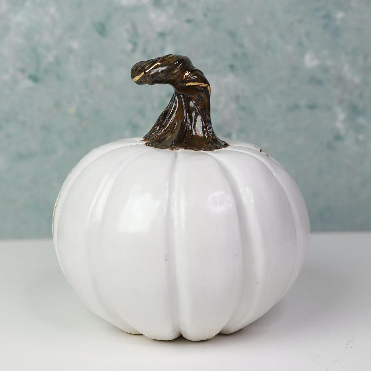 A glossy white decorative pumpkin figurine with a dark brown faux-stem topper, set against a textured light blue-gray background on a white surface.