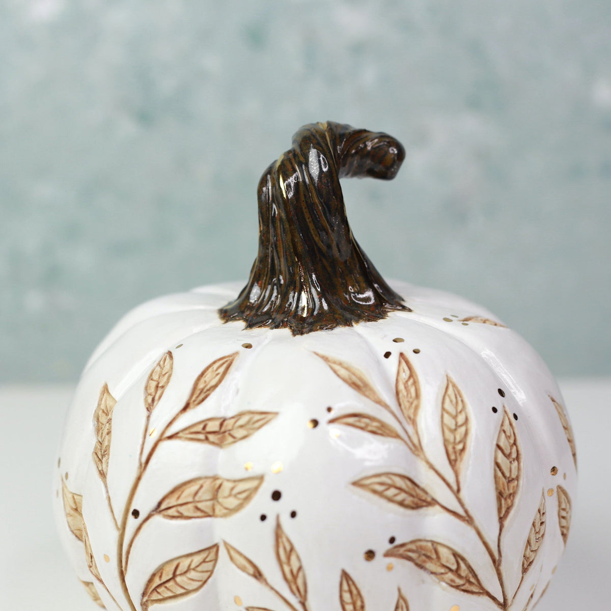 A white ceramic pumpkin figurine decorated with brown leafy vines and a dark brown textured stem, set against a soft, out-of-focus light blue-green background.