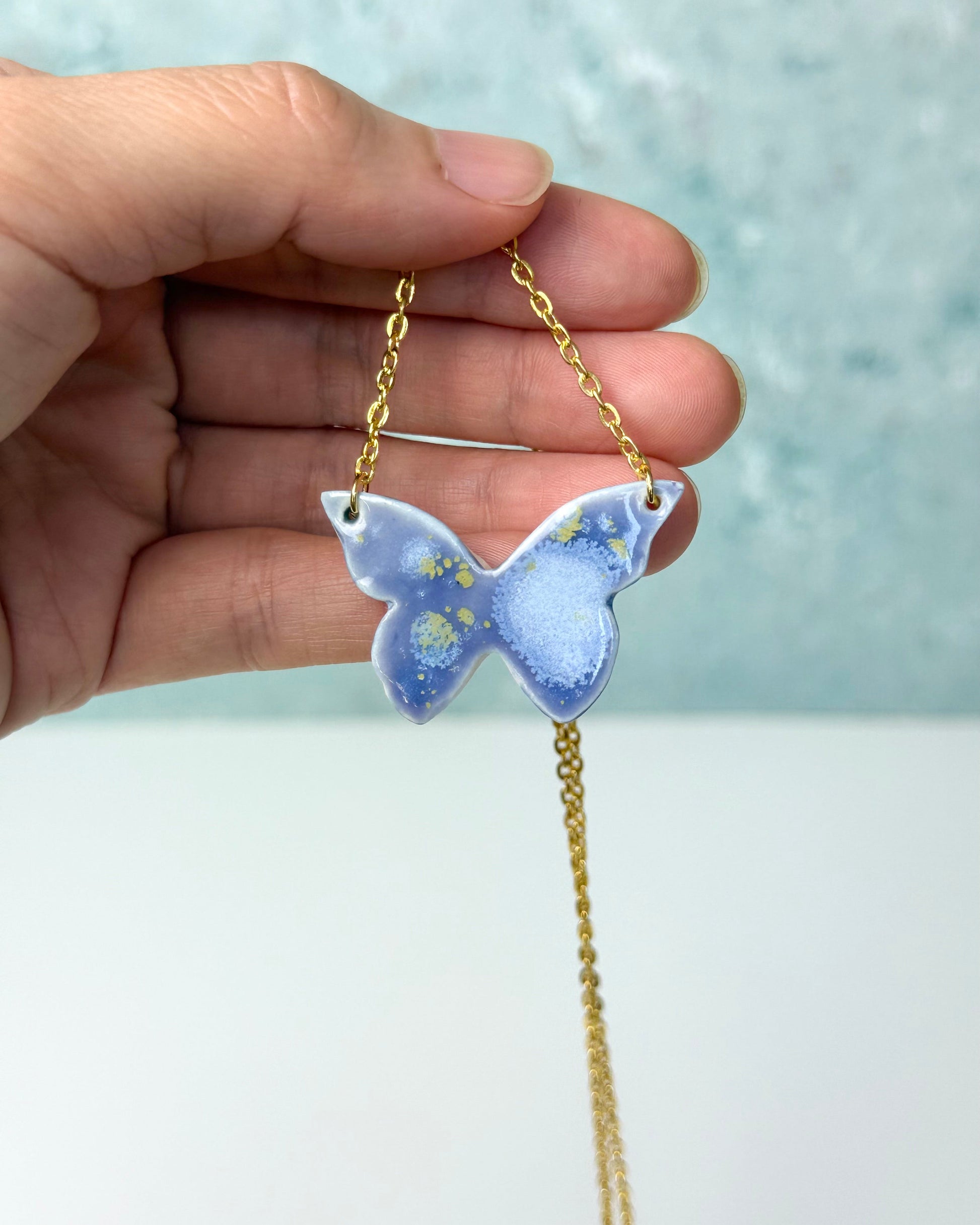 Butterfly-shaped pendant on a chain held by a hand against a light background