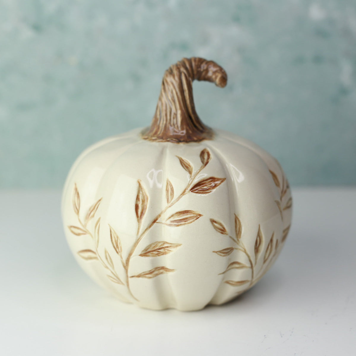 A decorative ceramic pumpkin figurine in cream white with brown leaf patterns and a brown stem, set against a soft, textured light blue-green background.