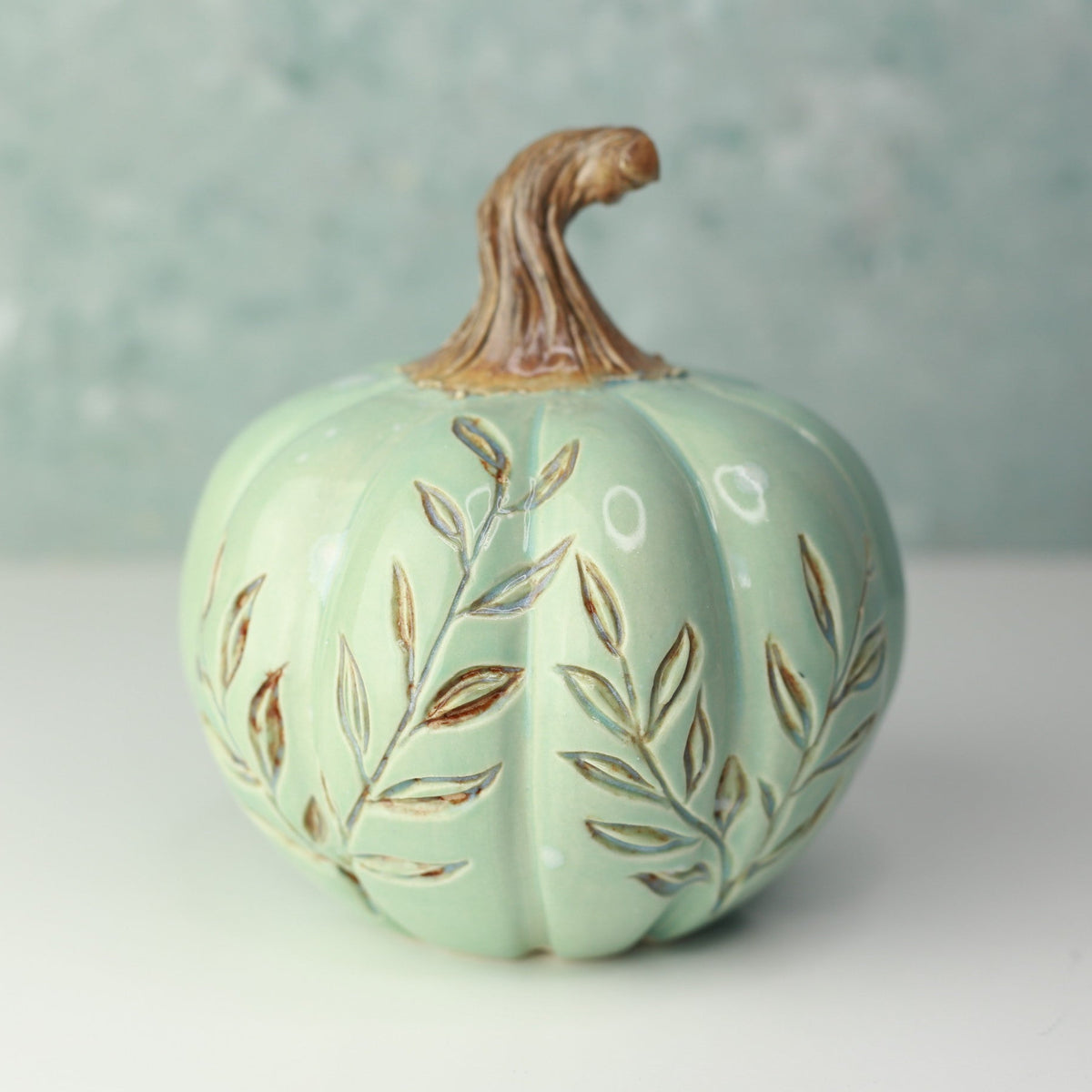 A decorative ceramic pumpkin in a pale mint green glaze with brown leafy vine patterns and a brown faux-stem top, shown on a white surface against a textured light blue-green background.