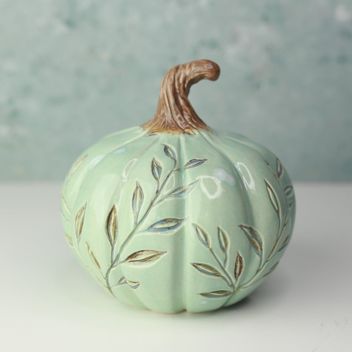 A decorative ceramic pumpkin in a pale mint green glaze with hand-painted gold and brown leaf patterns, featuring a realistic brown stem, set against a soft, textured light blue-gray background.