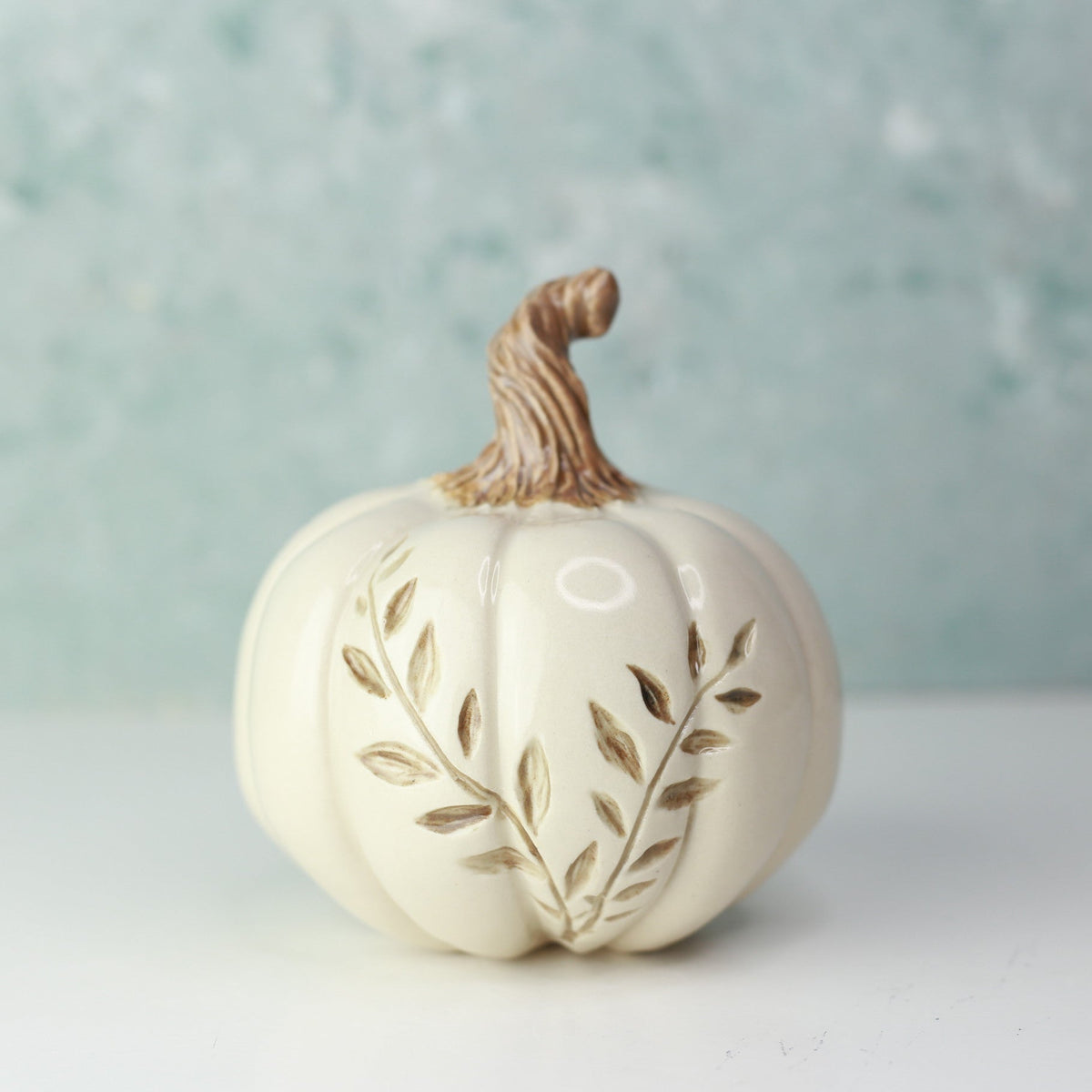 A small decorative ceramic pumpkin in warm off-white with a brown stem and embossed leaf details, set against a soft light blue textured background.