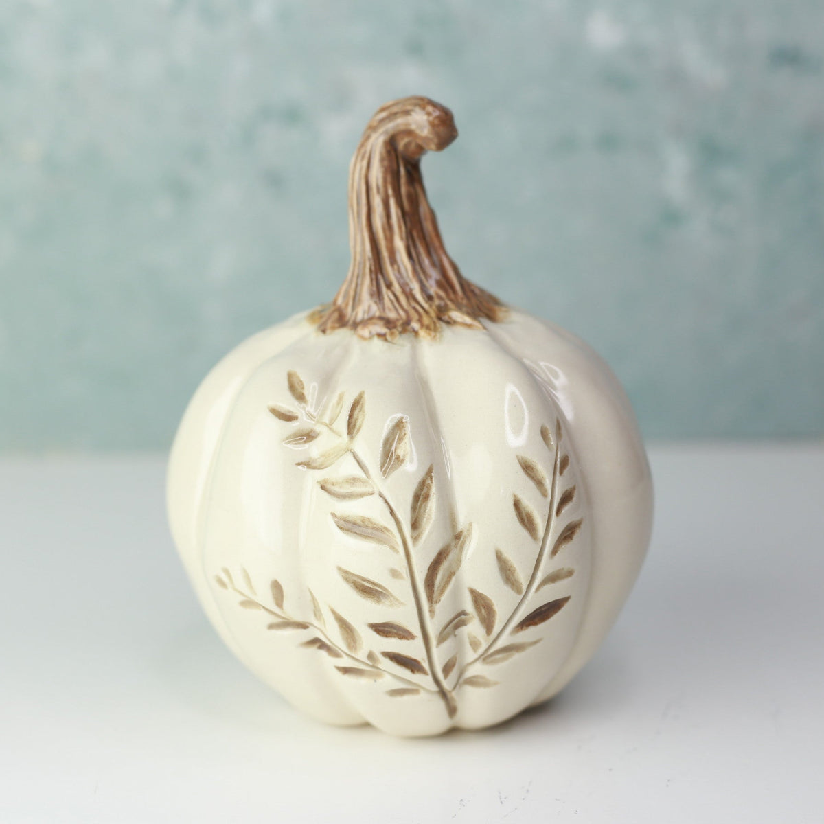 An off-white ceramic decorative pumpkin with a brown stem and embossed leaf details, set against a soft, textured light blue-green background.