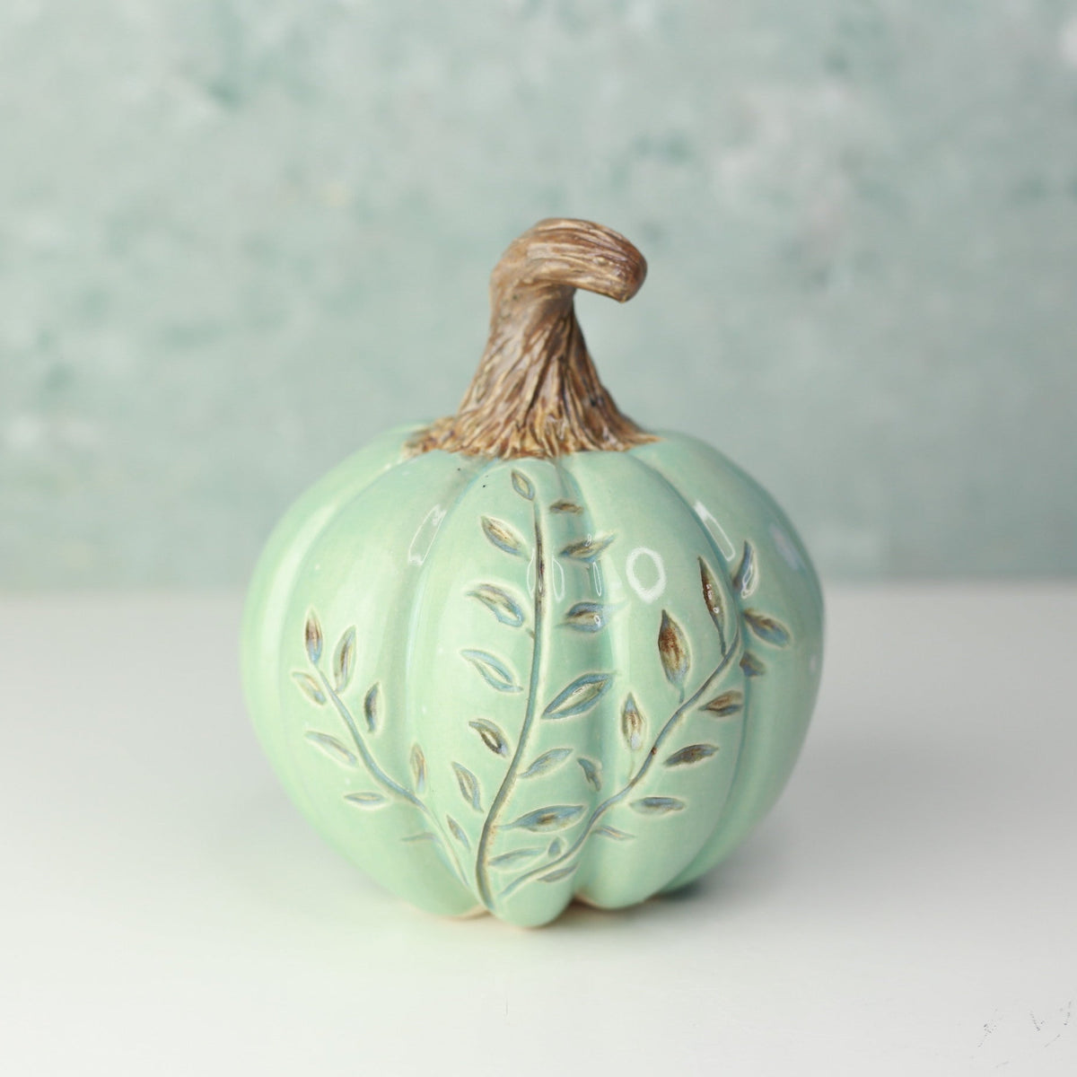 A small decorative ceramic pumpkin in a pale mint green color with leaf cutouts and a brown faux-stem top, set against a soft, textured light blue-green background.