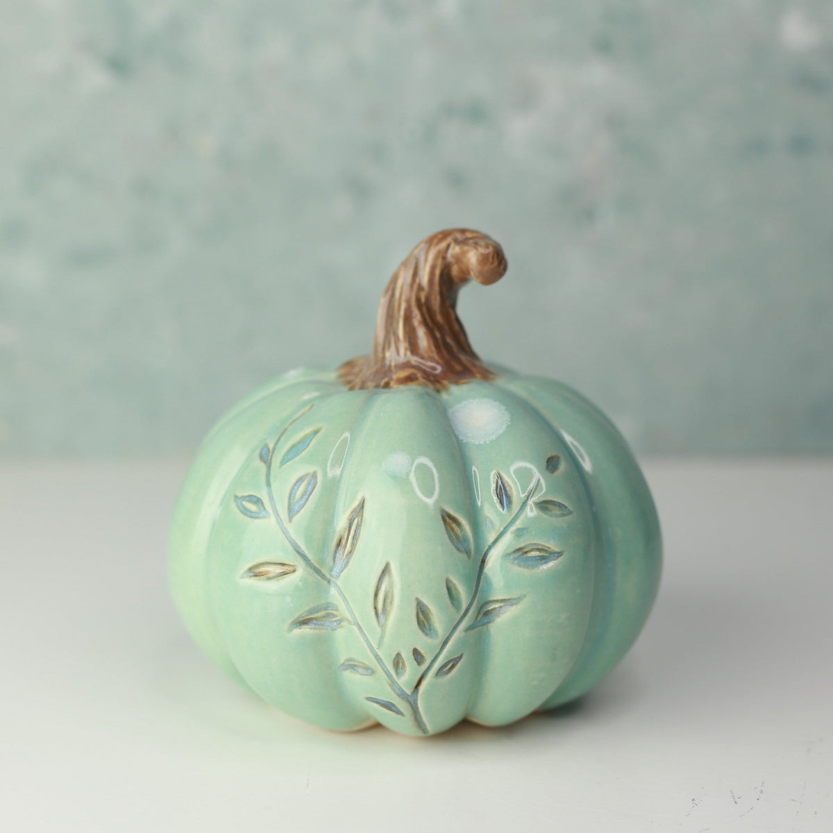 A small decorative ceramic pumpkin in mint green with a brown stem, featuring hand-painted gold and white leaf patterns on the front, set against a soft, textured light blue-green background.